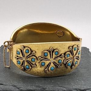 May include: A gold-tone hinged bangle bracelet featuring three raised floral motifs decorated with small turquoise-coloured cabochons and tiny white accents. The interior of the bracelet displays a circular stamp with the text FREIRICH.