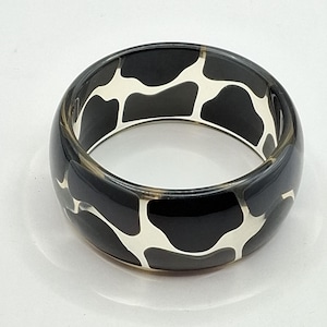Puede incluir: Una pulsera tipo brazalete ancha y circular con una base de resina gris oscuro brillante y translúcida, que presenta un patrón blanco opaco incrustado de formas orgánicas irregulares que envuelven toda la superficie exterior de la pieza.