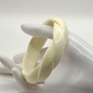 Könnte beinhalten: Elfenbeinfarbenes, gedrehtes Armreif-Armband. Das Armband hat eine glänzende Oberfläche und ein geflochtenes Design. Das Armband wird auf einem weißen, handförmigen Display gezeigt. Ein klassisches Accessoire.