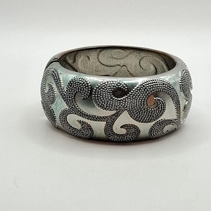May include: A wide, silver-toned hinged bangle bracelet featuring a raised, textured scrollwork pattern. The surface displays a contrast between smooth metallic areas and intricate, granular detailing within the swirling designs.