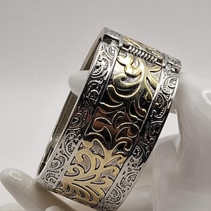 May include: A hinged bangle bracelet featuring a wide band with a two-tone finish. The centre section displays a raised gold-coloured floral scroll pattern against a dark background, flanked by silver-coloured borders with intricate engraved details.