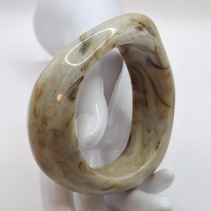 Peut inclure: Un bracelet jonc marbré dans les tons beige, marron et crème. Le bracelet a une forme carrée arrondie et est présenté sur une main blanche stylisée. La surface du bracelet a un aspect naturel et organique.