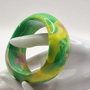 May include: A vibrant, marbled bangle bracelet in shades of green, yellow, and pink. The chunky, resin-like bracelet is displayed on a white hand, showcasing its colorful, swirled design. The bracelet's smooth, rounded edges and bold color palette make it a striking accessory.