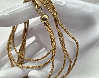 May include: A gold-colored necklace with multiple strands of braided chain. The necklace is draped over a white surface, showcasing its intricate design and the warm tones of the metal. The necklace has a clasp and two spherical end pieces.