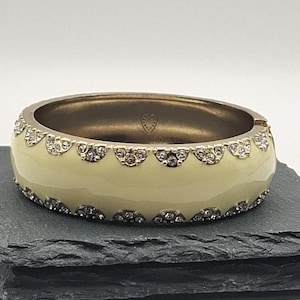 May include: Cream-coloured hinged bangle bracelet with a gold-tone metal frame. The bracelet features a row of small, clear crystals along the top and bottom edges. A small heart is visible on the inside of the bracelet.