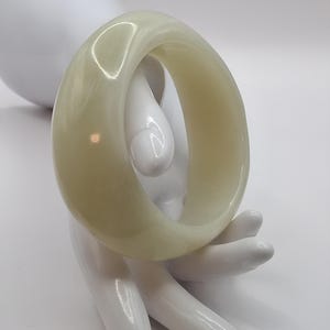Könnte beinhalten: Ein hellgrünes, durchscheinendes Armreif-Armband. Das glatte, abgerundete Armband wird auf einer weißen, stilisierten Handskulptur vor weißem Hintergrund präsentiert. Das Armband hat ein schlichtes, elegantes Design.