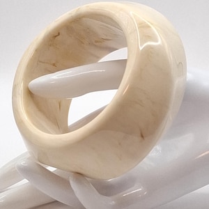 Op de afbeelding: Een crèmekleurige, brede armband met een gemarmerd effect. Het gladde, afgeronde ontwerp heeft een grote opening. De armband wordt getoond op een witte hand, wat de vorm en kleur benadrukt. Dit accessoire is geschikt voor dagelijks gebruik.
