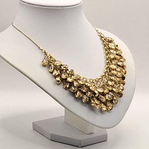 Op de afbeelding: Een goudkleurige statement ketting met meerdere lagen bungelende, druppelvormige bedels. De ketting is opgebouwd met een fijne kettingbasis en een dichte cluster van metalen, reflecterende hangers die een trapsgewijs, franjeachtig effect creëren.