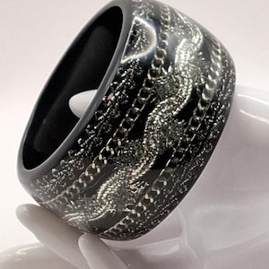 Op de afbeelding: Een brede, zwarte armband met een zilverkleurig, kettingachtig patroon. De armband is dik en heeft een glanzende afwerking. Het zilveren patroon is gedetailleerd en creëert een opvallend contrast met de zwarte basis.
