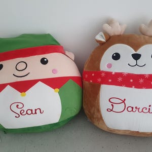 May include: Two festive plush cushions. One is an elf with a green hat and red trim, the other a reindeer with antlers and a red scarf. Each cushion has a name embroidered on it, perfect for the holiday season.