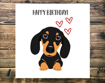 Puede incluir: Una tarjeta de felicitación con el texto "Happy Birthday!" y una imagen de dibujos animados de un dachshund negro y marrón con corazones rojos encima.