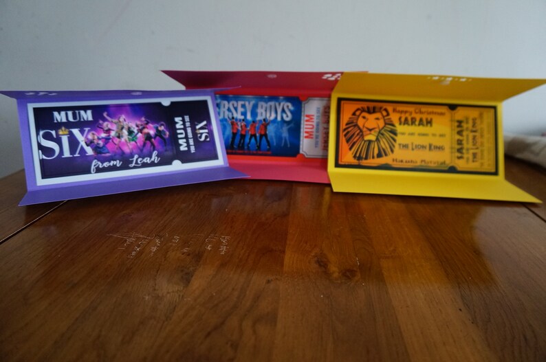 Puede incluir: Tres tarjetas plegadas con diferentes dise&ntilde;os. La primera tarjeta es morada y presenta una foto de un grupo musical con el texto "MUM SIX from Leah". La segunda tarjeta es roja y presenta una foto de un grupo musical con el texto "JERSEY BOYS". La tercera tarjeta es amarilla y presenta un le&oacute;n con el texto "Happy Christmas SARAH THE LION KING".