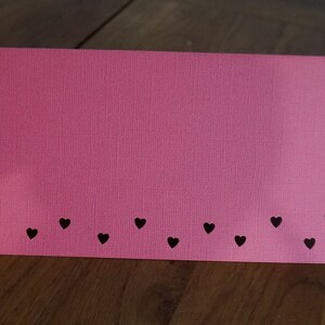 Puede incluir: Una tarjeta rosa con una fila de corazones negros a lo largo del borde inferior.