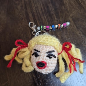 May include: A crocheted keychain featuring a doll head with blonde yarn hair styled in pigtails tied with red yarn bows. The face has black embroidered eyebrows, black bead eyes, and red lips. A silver metal clasp is attached to a string of colorful letter beads.