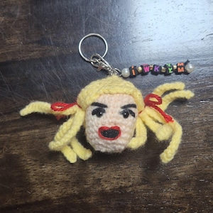 May include: A handmade crochet keychain featuring a face with blonde hair, red bows, and a red mouth. The keychain includes a silver ring and a beaded charm spelling out "I W K Y M".
