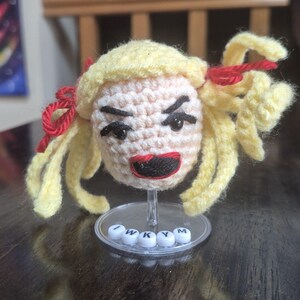 May include: Crocheted doll head with yellow hair, red bows, and a stern expression. The doll has black eyes and a red mouth. The base reads "I W K Y M".