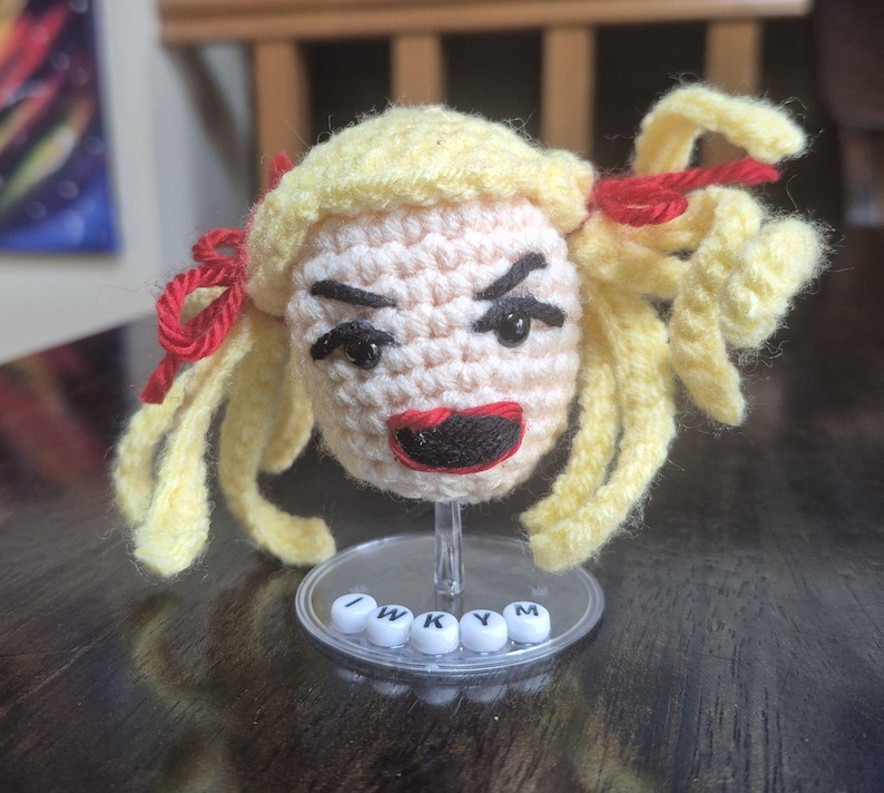 May include: Crocheted doll head with yellow hair, red bows, and a stern expression. The doll has black eyes and a red mouth. The base reads "I W K Y M".