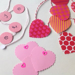 Può includere: Una ghirlanda di cuori di carta rosa e rossa con un motivo a pois rosa e bianco. La ghirlanda è infilata su uno spago bianco e ha cerchi di carta rosa con le lettere "i", "e", "j", "f" e "e" scritte in nero.