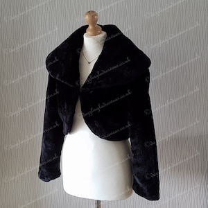 May include: A black faux fur bolero jacket with a wide collar. The jacket is cropped at the waist and has long sleeves.