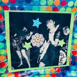 May include: A black and white photo of a band playing music. The photo is framed with a green border and decorated with colorful stickers, including a smiley face flower and stars. The band's name, "GLES", is visible on a drum.