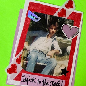 May include: A pink and red card with a photo of a young man sitting in a chair. The card has a black star, pink heart, and red hearts with white stitching. The text on the card reads "Back to the Cave!"