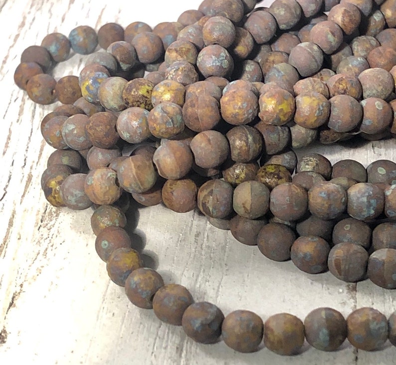 May include: Strands of round, matte beads in shades of brown, with hints of blue and yellow. The beads have a rustic, weathered appearance, suggesting a natural or vintage aesthetic. Ideal for jewelry making or craft projects.
