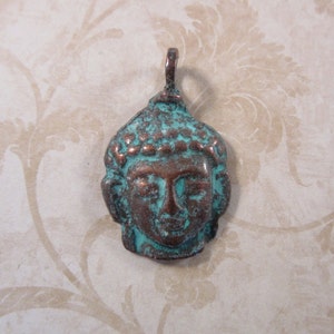 May include: A weathered bronze-colored pendant in the shape of a Buddha's head. The pendant has a turquoise patina on the surface, with visible details of the face and hair. The pendant has a loop at the top for hanging.