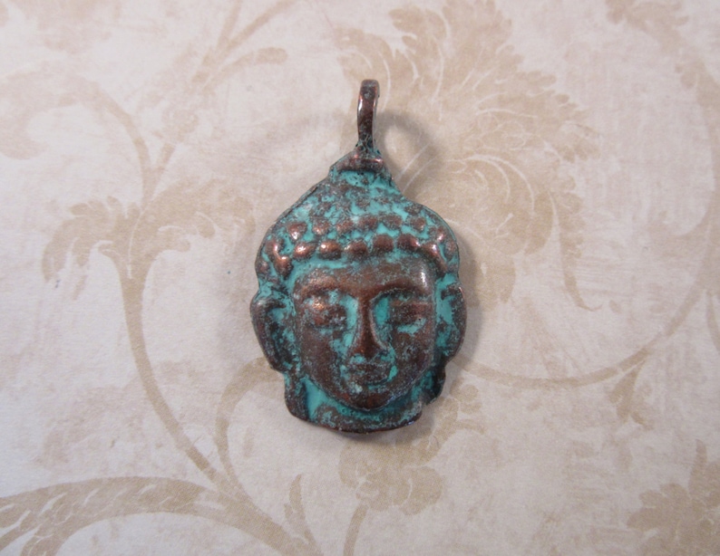 May include: A weathered bronze-colored pendant in the shape of a Buddha's head. The pendant has a turquoise patina on the surface, with visible details of the face and hair. The pendant has a loop at the top for hanging.