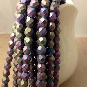May include: Strands of iridescent faceted beads in shades of purple, green, and blue. The beads are draped over a white ceramic vase. The beads have a geometric, multi-faceted design.