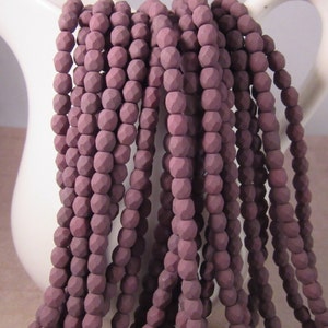 May include: Strands of mauve-colored faceted beads cascade from a white pitcher. The beads have a matte finish and a geometric, multi-faceted design. The beads are strung together, creating a flowing, textured appearance, suitable for jewelry making.