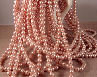 May include: A collection of blush pink pearl necklaces cascading from a white ceramic pitcher. The pearls are uniform in size and have a subtle sheen. The necklaces are draped and arranged to create a visually appealing display.