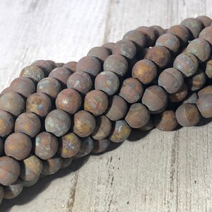 May include: Close-up of a strand of round, weathered beads. The beads have a mottled appearance with shades of brown, bronze, and hints of blue and yellow. The beads are likely used for jewelry making.