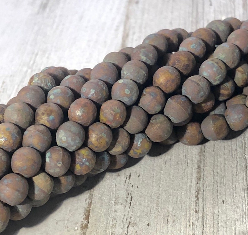 May include: Close-up of a strand of round, weathered beads. The beads have a mottled appearance with shades of brown, bronze, and hints of blue and yellow. The beads are likely used for jewelry making.