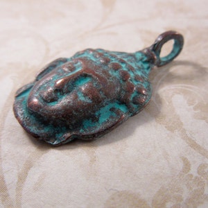 May include: A weathered metal pendant in the shape of a Buddha head. The pendant has a copper-colored base with turquoise-colored accents, giving it an aged appearance. The pendant has a loop at the top for hanging.