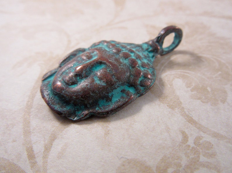 May include: A weathered metal pendant in the shape of a Buddha head. The pendant has a copper-colored base with turquoise-colored accents, giving it an aged appearance. The pendant has a loop at the top for hanging.