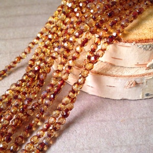 May include: Strands of faceted amber-colored beads with a mix of reddish-brown hues. The beads are strung together, creating a shimmering effect. The beads are displayed on a light-colored surface.