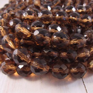 May include: Close-up of a strand of faceted, dark brown beads. The beads have a multifaceted design, catching light and reflecting a warm, amber hue. The beads are strung together, suggesting their use in jewelry or craft projects.