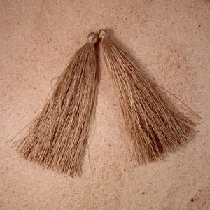 May include: Two beige-colored tassels, each with a looped top and a long, flowing fringe. The tassels are made of a silky, shiny material and are lying on a light brown, textured surface. These tassels could be used for jewelry making or as decorative accents.