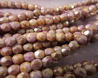 May include: Close-up of strands of faceted beads in shades of mauve, tan, and gold. The beads have a slightly irregular shape and a subtle sheen, suggesting a vintage or handmade quality. The beads are strung together, creating a flowing, textured surface.
