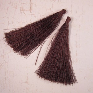May include: Two dark brown tassels with a silky texture. The tassels are made of many strands of thread, and each has a knotted top. The tassels are lying on a light-colored surface.