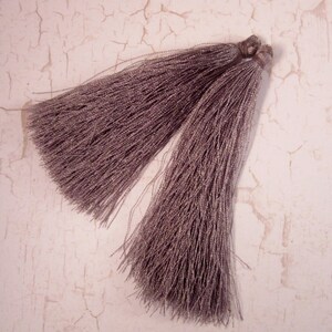May include: Two mauve-colored tassels with a silky, shimmering appearance. The tassels are made of many fine strands, tied together at the top. The background is a light, textured surface with a cracked, aged look.