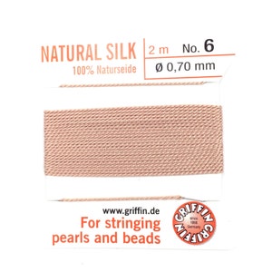 May include: A spool of natural silk thread, color peach, for stringing pearls and beads. The label reads "NATURAL SILK" and "100% Naturseide". The thread is 2 meters long with a diameter of 0.70 mm. The brand is Griffin, since 1866, Germany.