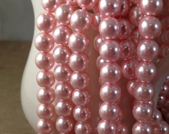 May include: Strands of pink faux pearls cascade from a white ceramic pitcher. The pearls are a soft, rosy hue with a subtle sheen, creating a classic and elegant look. The pearls are strung together, ready for jewelry making or other craft projects.