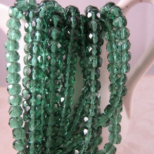 May include: Strands of faceted, emerald green beads. The beads are strung together, creating a shimmering effect. The beads are displayed in a white ceramic container. The beads are a deep, rich green color.
