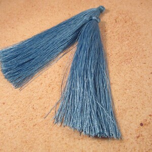 May include: Two blue-green tassels, likely for crafting or embellishment, lie side-by-side against a textured, light brown surface. The tassels are made of many fine, silky threads, creating a soft, flowing appearance. The color is a muted, dusty teal.