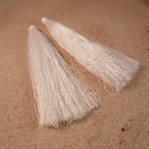 May include: Two white silk tassels, each with a looped top and a long, flowing fringe. The tassels are lying on a light brown, speckled surface. These decorative tassels are suitable for various craft projects.