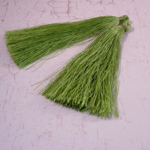 May include: Two bright green tassels made of fine, shiny threads. The tassels are tied at the top with a matching green thread, creating a decorative element. The tassels are lying on a light-colored surface.
