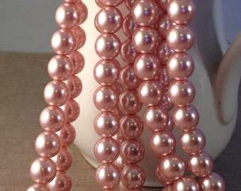 May include: Strands of pink faux pearls hang in front of a white ceramic pitcher. The pearls are a soft, rosy color and have a glossy finish. The pearls are strung together, creating a cascading effect.