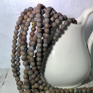 May include: Strands of brown, round beads with a textured, weathered appearance cascade from a white ceramic pitcher. The beads have a mix of brown, blue, and yellow tones, suggesting an aged or antique aesthetic. The beads are likely for jewelry making.