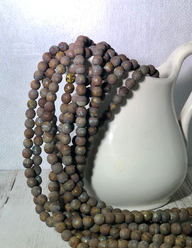 May include: Strands of brown, round beads with a textured, weathered appearance cascade from a white ceramic pitcher. The beads have a mix of brown, blue, and yellow tones, suggesting an aged or antique aesthetic. The beads are likely for jewelry making.